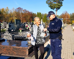 Policjant w mundurze daje ulotkę kobiecie sprzątającej na cmentarzu.