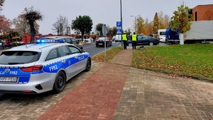 Radiowóz stojący na poboczu a policjant i strażnik miejski idą chodnikiem.