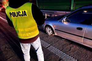 Policjant w kamizelce z napisem policja przy aucie w którym  widać brak powietrza w kole i klucz do odkręcania śruby.