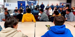 Prowadzący spotkanie policjanci, strażak i inne osoby stoją przed młodzieżą siedzącą w ławkach.