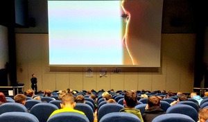 Na ekranie wyświetlany film, na którym widać twarz kobiety, na uli siedzą osoby przed  nimi stoi policjantka w mundurze.
