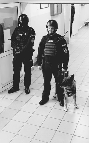 Dwóch policjantów, jeden z psem służbowym drugi z długa bronią w ręce.