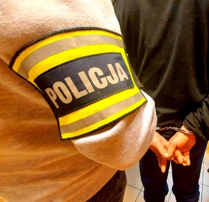 Opaska na ramieniu policjanta z napisem policja i ręce w kajdankach zatrzymanego.