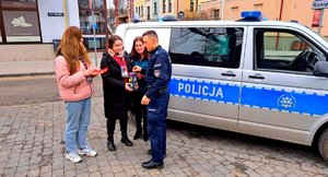 Policjant przy radiowozie wrzuca do skarbonki pieniądze obok 3 wolontariuszki.