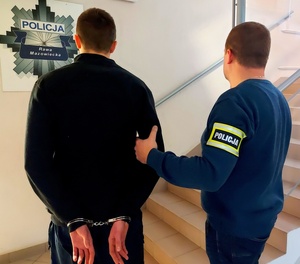 policjant w cywilnych ubraniach z opaską na ręce z napisem policja trzyma za przedramię zatrzymanego mężczyznę, który ma kajdanki na rękach założone z tyłu. Na ścianie widać gwiazdę policyjna z napisem Komenda Powiatowa Policji w Rawie Mazowieckiej.