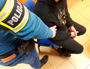 zatrzymany siedzący na krzesełku, na rękach kajdanki obok stoi policjant na ramieniu opaska a napisem policja.
