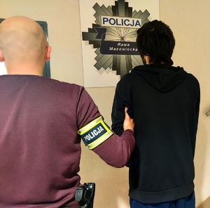 policjant w ubraniu cywilnym z opaska na ręce z napisem policja trzyma za rękę zatrzymanego mężczyznę, na ścianie gwiazda policyjna.
