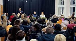 policjantka stoi z mikrofonem przed rodzicami, którzy siedzą w ławkach. komendant oraz dyrektorka siedzą.