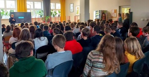 policjantka stojąca przy tablicy i dzieci siedzące w ławeczkach.