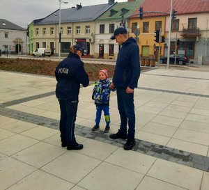 policjantka dająca opaskę odblaskową dziecku, które idzie z rodzicem za rękę.
