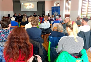seniorzy siedzący na krzesełkach tyłem do fotografa a przed nimi policjanci w mundurach i na ścianie wyświetla się film.