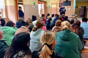 policjant stojący przed rodzicami siedzącymi na ławeczkach.