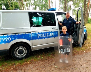 Policjant stojący przy radiowozie z małym chłopcem, który trzyma w ręce tarczę policyjną i pałkę służbową.