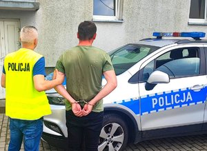 Policjant w kamizelce z napisem policja trzyma za rękę mężczyznę, który ma założone kajdanki. Stoją obok radiowozu.