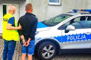 Policjant w kamizelce z napisem policja trzyma za rękę mężczyznę w krótkich spodenkach, który ma założone kajdanki. Stoją obok radiowozu.