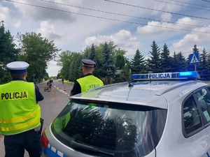 Policjant i strażnik miejski stoją obok radiowozu, na drodze jedzie na rowerze uczestnik zawodów.