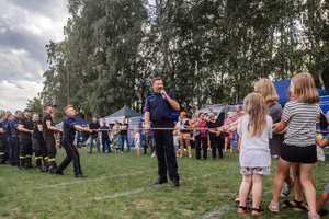 Policjanci trzymają za linę, pośrodku Komendant z mikrofonem a po drugiej stronie dzieci.