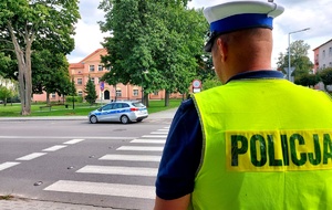 Policjant stojący tyłem, przed nim przejście dla pieszych, radiowóz i szkoła.