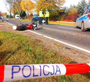 Policjanci stojący przy aucie, leżący motocykl na jezdni i taśma z napisem policja.