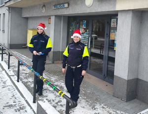 Policjantki w czapkach Mikołaja zawieszają odblaski na poręczy przed komendą.