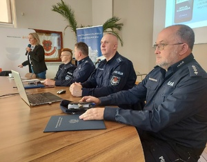 Policjanci siedzące przy stole i kobieta z mikrofonem.