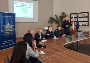 Policjant stojący z mikrofonem, inni policjanci siedzący i inne osoby.