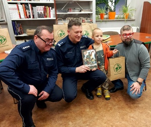 Policjanci oraz dyrektor bibliotetki kucają obok chłopca z nagrodą w ręce.