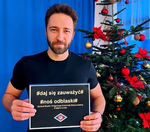 Mężczyzna trzymający kartkę w ręce z napisem daj się zauważyć noś odblaski obok choinka.