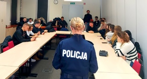 Policjantka stojąca tyłem i młodzież siedząca przy stole.