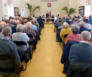 Osoby siedzące na krzesłach i w oddali z mikrofonem policjantka.