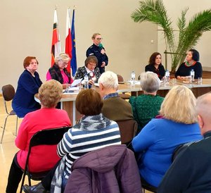 Policjantka stojąca z mikrofonem i seniorzy siedzący na krzesłach.