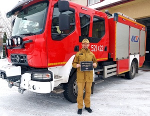 Komendant Straży Pożarnej stojący przy radiowozie  z kartką z napisem daj się zauważyć noś odblaski.