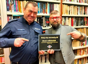 Policjant i dyrektor biblioteki z kartką z napisem daj się zauważyć noś odblaski na tle regałów z książkami.