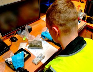 Policjant w kamizelce siedzący przy biurku na którym widać wagę elektroniczną, dokumenty, woreczki z narkotykami.