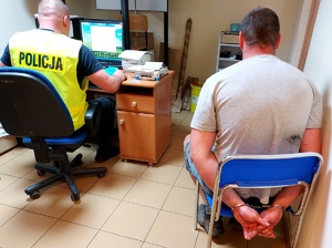 Mężczyzna z założonymi kajdankami na rękach siedzący na krzesełku i policjant siedzący przy komputerze.