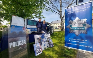Policjanci stoją przy stoliku z wystawionym sprzętem za nimi radiowóz.