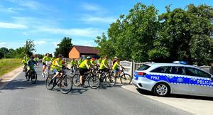 Osoby jadące na rowerach w kamizelkach odblaskowych za radiowozem.