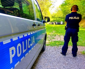 Radiowóz z napisem policja i stojący policjant w tle tarcza do strzelania.
