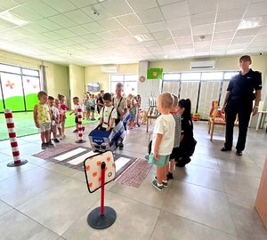 Jedna policjantka stoi, druga kuca przy dzieciach, które uczą się przechodzić po macie edukacyjnej w przez przejście dla pieszych.