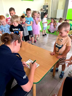 Policjantka uzupełnia certyfikat dla przedszkolaka, dzieci stoją w kolejce.