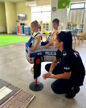 Policjantka kucająca przy dzieciach, dzieci ubrane w autochodzik.