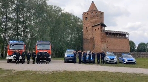 Radiowozy straży pożarnej oraz policji, przed nimi strażacy i policjanci salutują, na tle ruin zamku rawskiego.