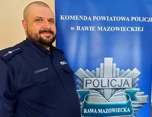 Policjant stojący na tle napisu Komenda Powiatowa Policji w Rawie Mazowieckiej.