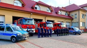 Radiowozy policyjne oraz straży pożarnej z włączonymi sygnałami błyskowymi oraz policjanci i strażacy na baczność.