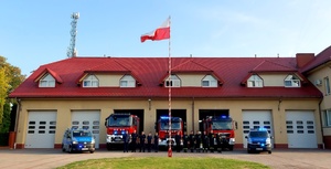 Budynek straży pożarnej, radiowozy policyjne i strażackie oraz policjanci i strażacy oddający cześć na baczność.