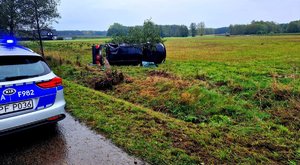 Radiowóz oraz samochód leżący na boku na polu i przewrócone drzewo.