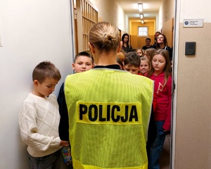 Policjantka w kamizelce z napisem policja stojąca tyłem przed nią dzieci.