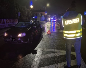 Policjant w kamizelce z napisem policja, wykonujący zdjęcia na miejscu wypadku, obok samochód na drodze,