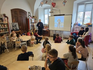 Policjant, dzieci siedzące przy stolikach i na ekranie zdjęcie policjanta z psem.