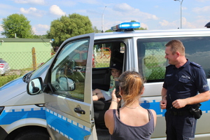 Policjant przy radiowozie obok stojące inne osoby.